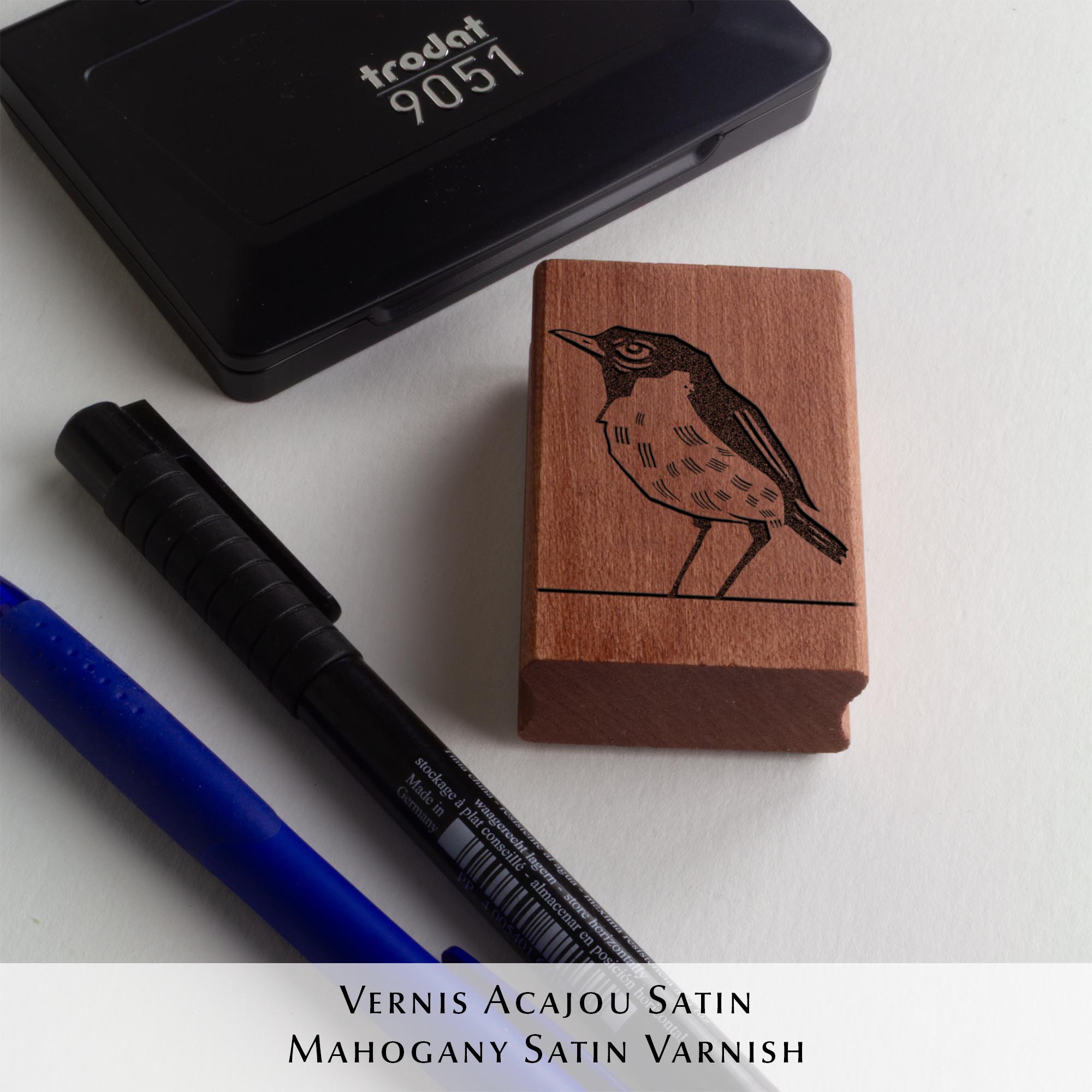 Deux tampons Ex-Libris en bois gravés d'un oiseau, présentés côte à côte au creux de deux mains. Des lignes de texte indiquent la différence de finition : à gauche, le bois a une teinte rousse avec la mention "Vernis Acajou Satin", et à droite, une teinte naturelle claire avec la mention "Vernis Incolore Brillant".