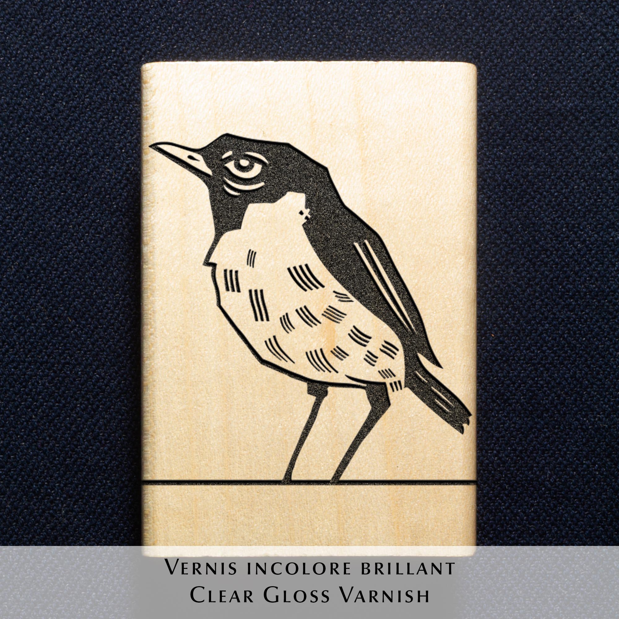 Deux tampons Ex-Libris en bois gravés d'un oiseau, présentés côte à côte au creux de deux mains. Des lignes de texte indiquent la différence de finition : à gauche, le bois a une teinte rousse avec la mention "Vernis Acajou Satin", et à droite, une teinte naturelle claire avec la mention "Vernis Incolore Brillant".