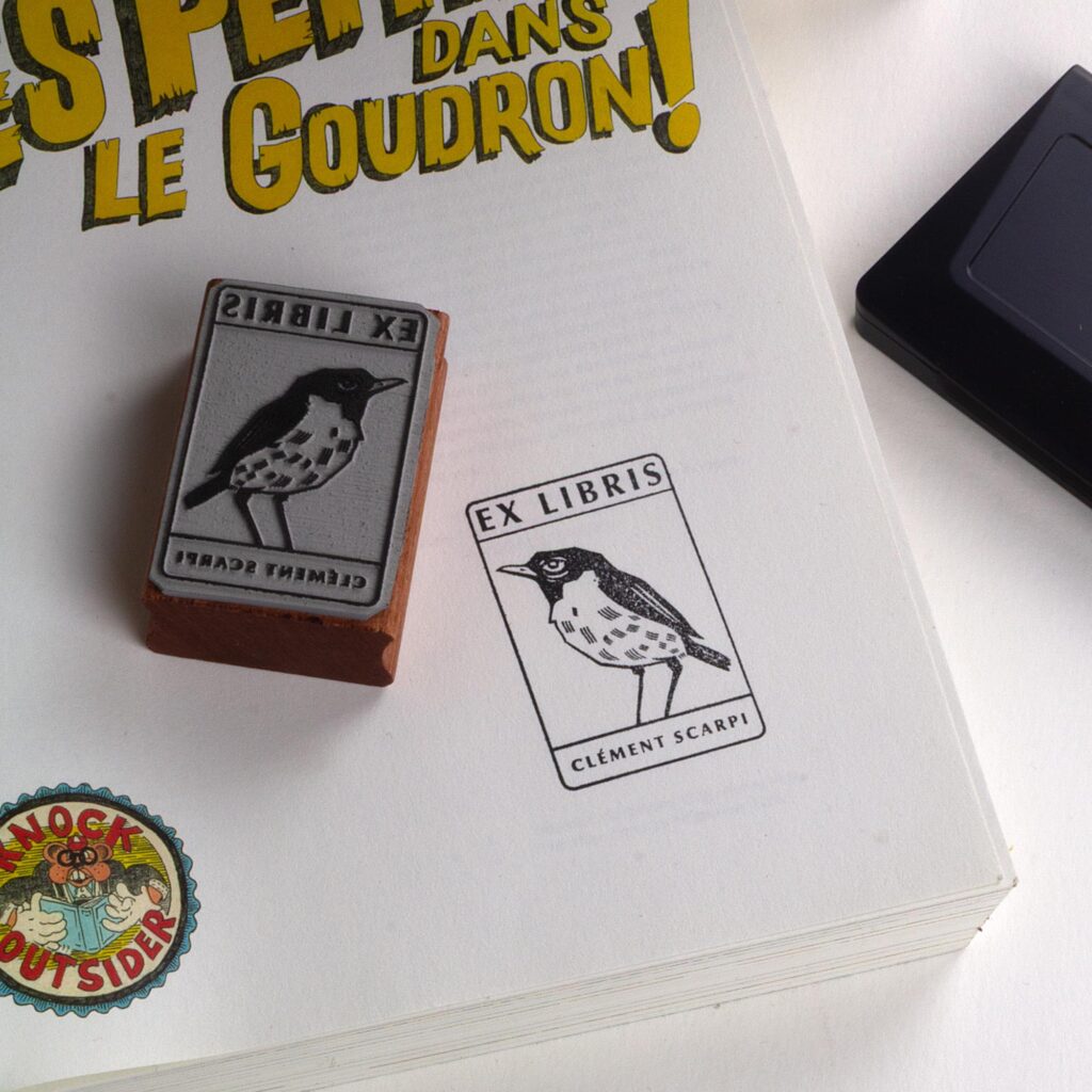 Deux tampons Ex-Libris en bois gravés d'un oiseau, présentés côte à côte au creux de deux mains. Des lignes de texte indiquent la différence de finition : à gauche, le bois a une teinte rousse avec la mention "Vernis Acajou Satin", et à droite, une teinte naturelle claire avec la mention "Vernis Incolore Brillant".