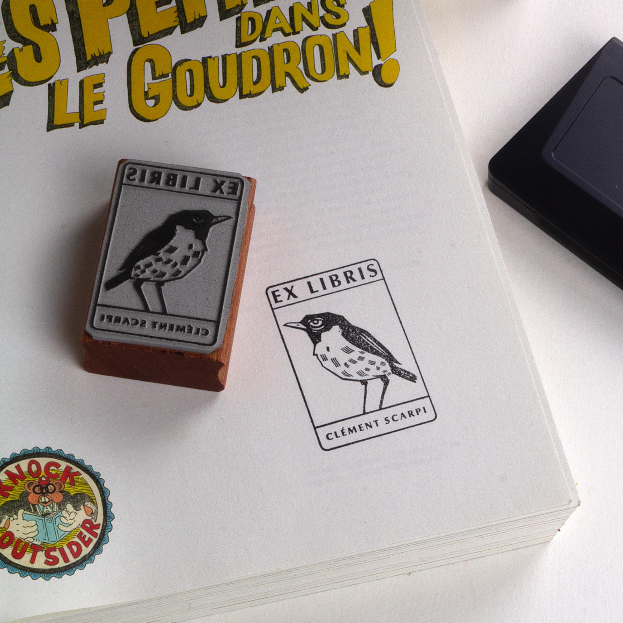 Deux tampons Ex-Libris en bois gravés d'un oiseau, présentés côte à côte au creux de deux mains. Des lignes de texte indiquent la différence de finition : à gauche, le bois a une teinte rousse avec la mention "Vernis Acajou Satin", et à droite, une teinte naturelle claire avec la mention "Vernis Incolore Brillant".