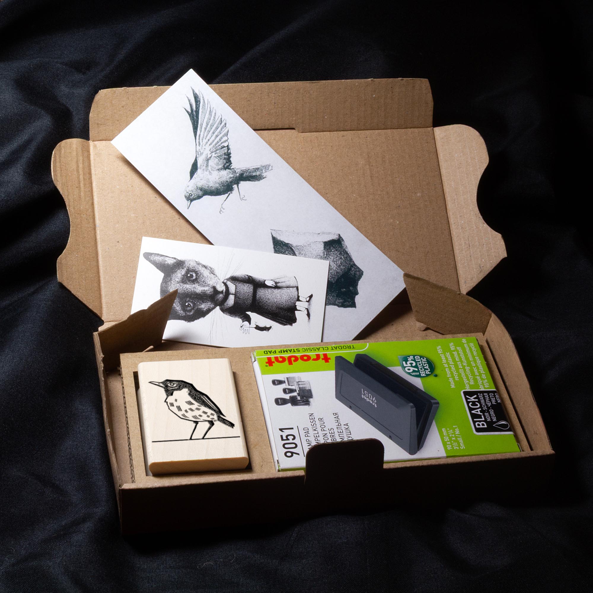 Deux tampons Ex-Libris en bois gravés d'un oiseau, présentés côte à côte au creux de deux mains. Des lignes de texte indiquent la différence de finition : à gauche, le bois a une teinte rousse avec la mention "Vernis Acajou Satin", et à droite, une teinte naturelle claire avec la mention "Vernis Incolore Brillant".