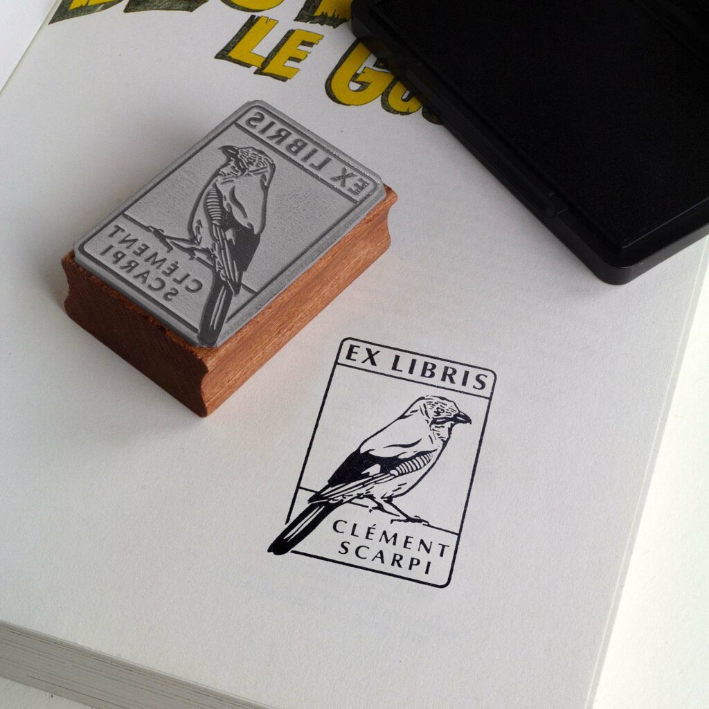 Deux tampons Ex-Libris en bois gravés d'un geai des chênes, présentés côte à côte au creux de deux mains. Des lignes de texte indiquent la différence de finition : à gauche, le bois a une teinte rousse avec la mention "Vernis Acajou Satin", et à droite, une teinte naturelle claire avec la mention "Vernis Incolore Brillant".
