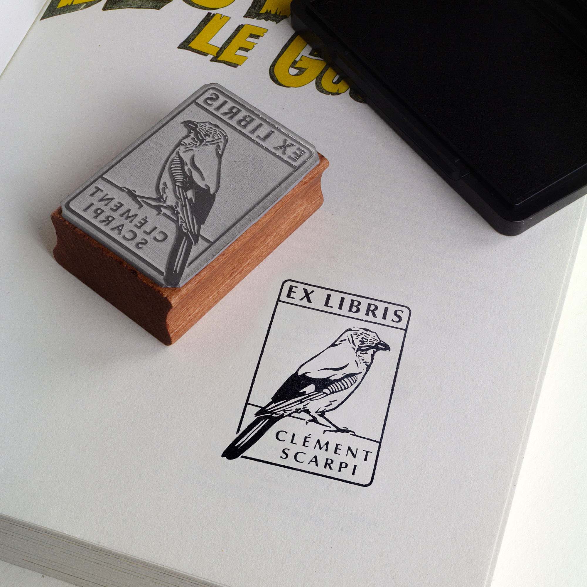 Deux tampons Ex-Libris en bois gravés d'un geai des chênes, présentés côte à côte au creux de deux mains. Des lignes de texte indiquent la différence de finition : à gauche, le bois a une teinte rousse avec la mention "Vernis Acajou Satin", et à droite, une teinte naturelle claire avec la mention "Vernis Incolore Brillant".