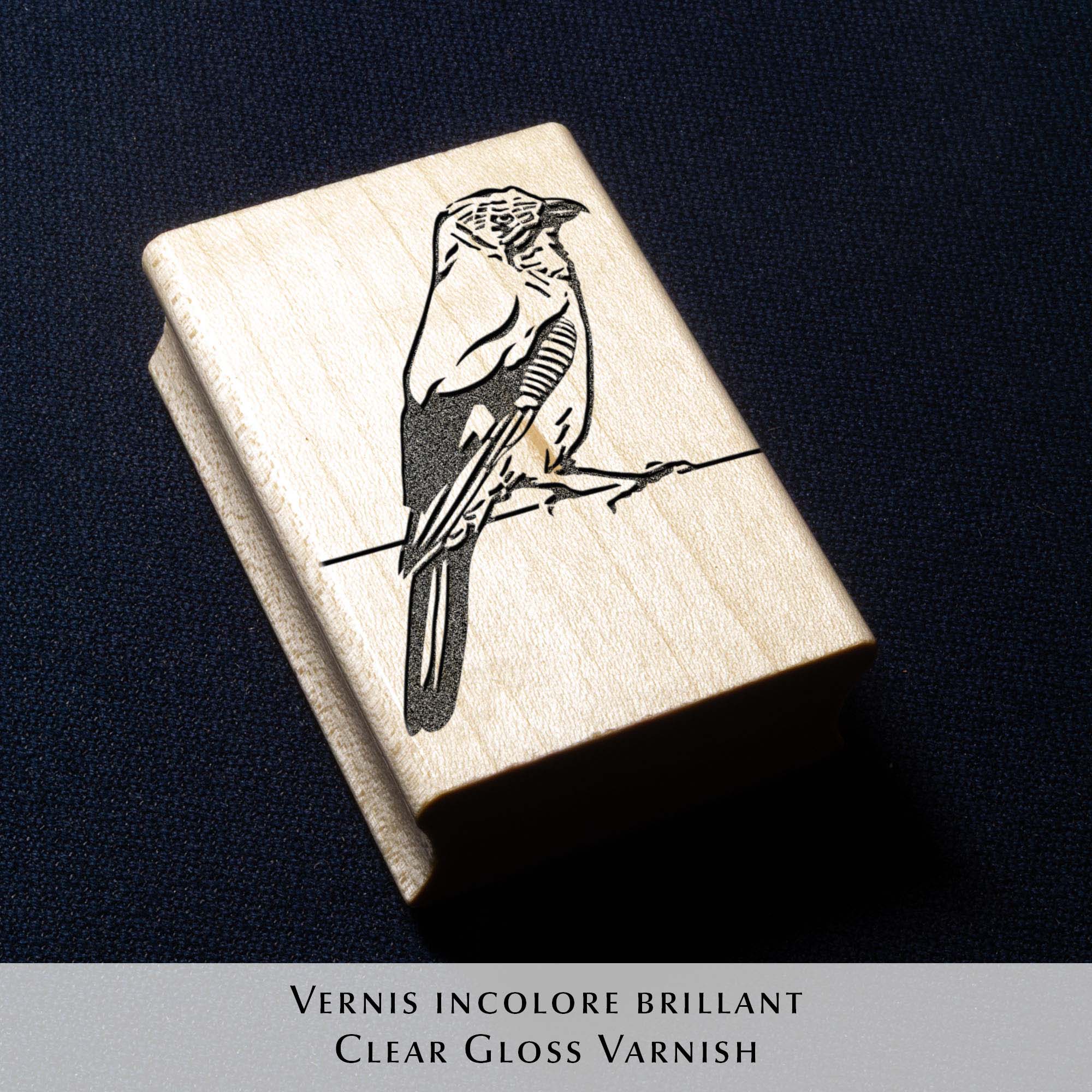 Deux tampons Ex-Libris en bois gravés d'un geai des chênes, présentés côte à côte au creux de deux mains. Des lignes de texte indiquent la différence de finition : à gauche, le bois a une teinte rousse avec la mention "Vernis Acajou Satin", et à droite, une teinte naturelle claire avec la mention "Vernis Incolore Brillant".