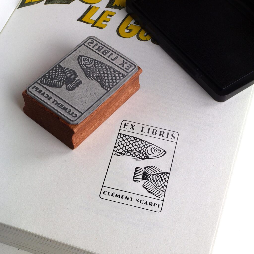 Deux tampons Ex-Libris en bois gravés d'un poisson, présentés côte à côte au creux de deux mains. Des lignes de texte indiquent la différence de finition : à gauche, le bois a une teinte rousse avec la mention "Vernis Acajou Satin", et à droite, une teinte naturelle claire avec la mention "Vernis Incolore Brillant".