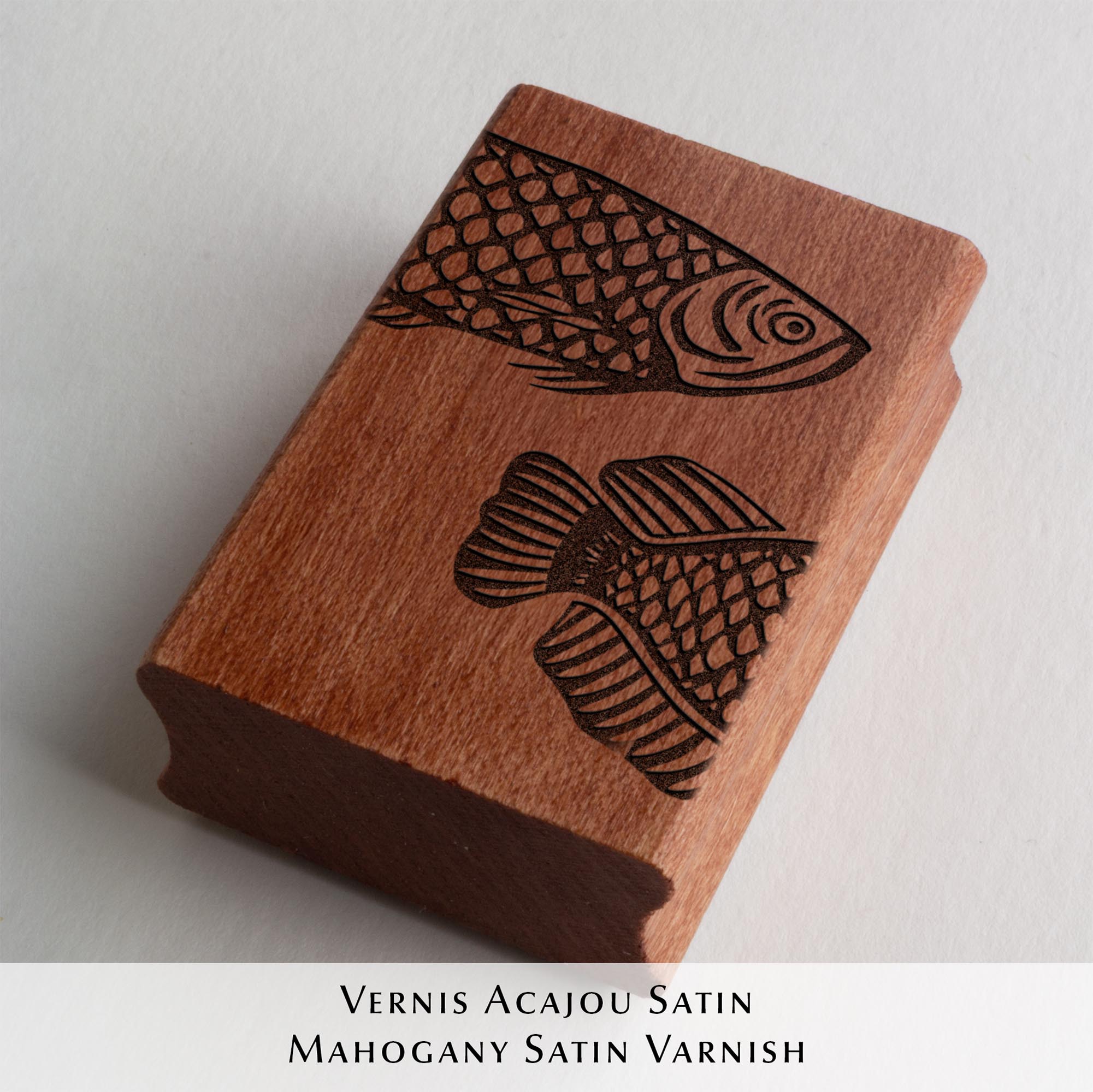 Deux tampons Ex-Libris en bois gravés d'un poisson, présentés côte à côte au creux de deux mains. Des lignes de texte indiquent la différence de finition : à gauche, le bois a une teinte rousse avec la mention "Vernis Acajou Satin", et à droite, une teinte naturelle claire avec la mention "Vernis Incolore Brillant".