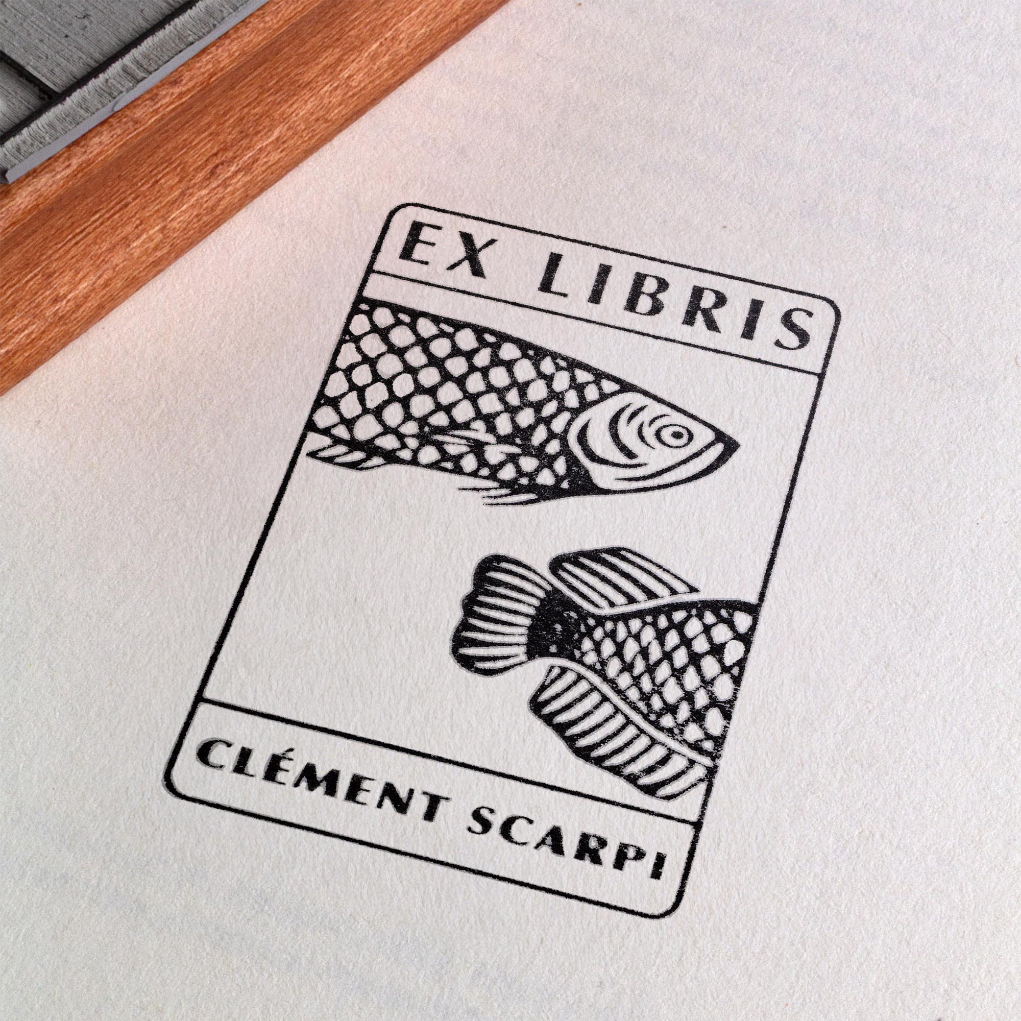 Deux tampons Ex-Libris en bois gravés d'un poisson, présentés côte à côte au creux de deux mains. Des lignes de texte indiquent la différence de finition : à gauche, le bois a une teinte rousse avec la mention "Vernis Acajou Satin", et à droite, une teinte naturelle claire avec la mention "Vernis Incolore Brillant".