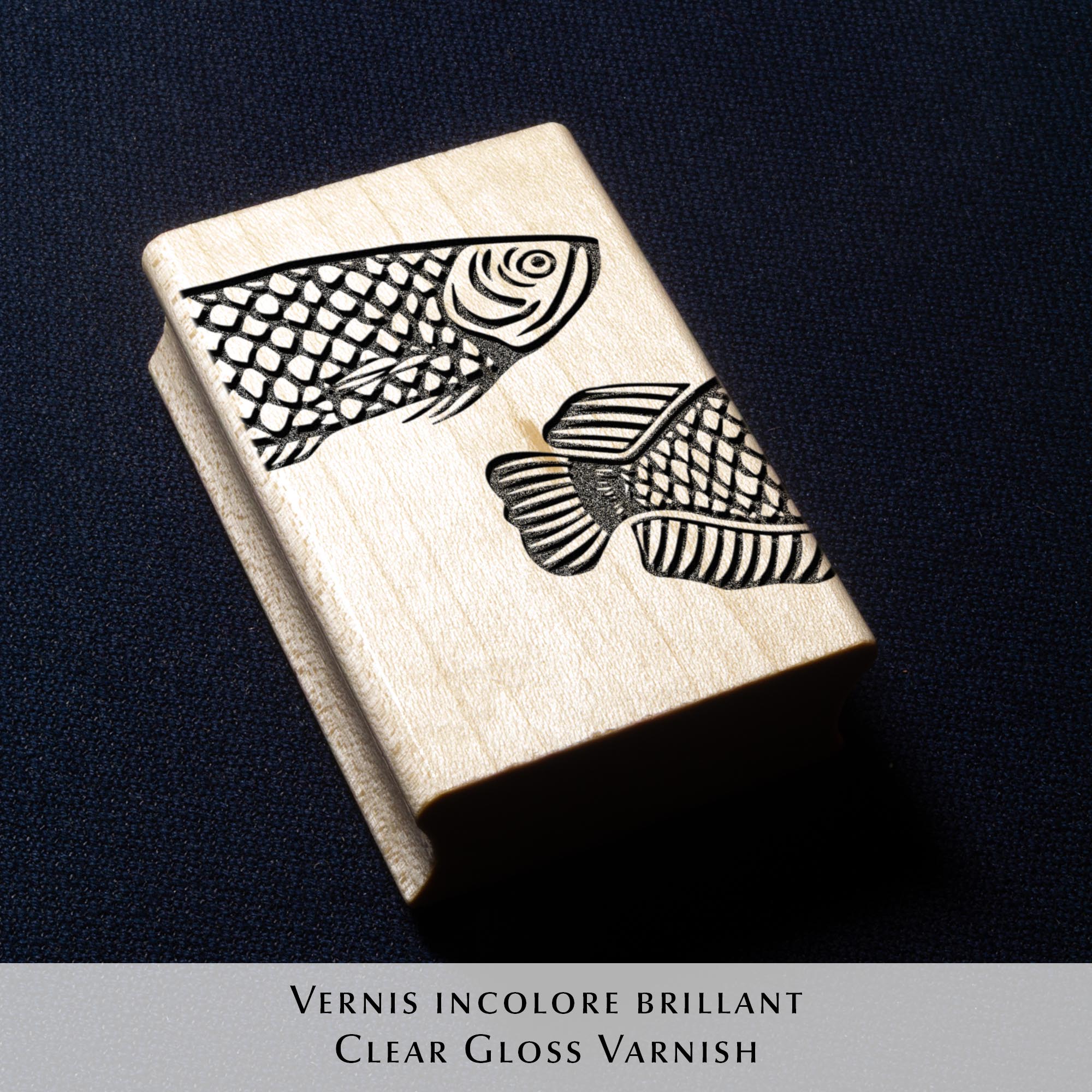 Deux tampons Ex-Libris en bois gravés d'un poisson, présentés côte à côte au creux de deux mains. Des lignes de texte indiquent la différence de finition : à gauche, le bois a une teinte rousse avec la mention "Vernis Acajou Satin", et à droite, une teinte naturelle claire avec la mention "Vernis Incolore Brillant".