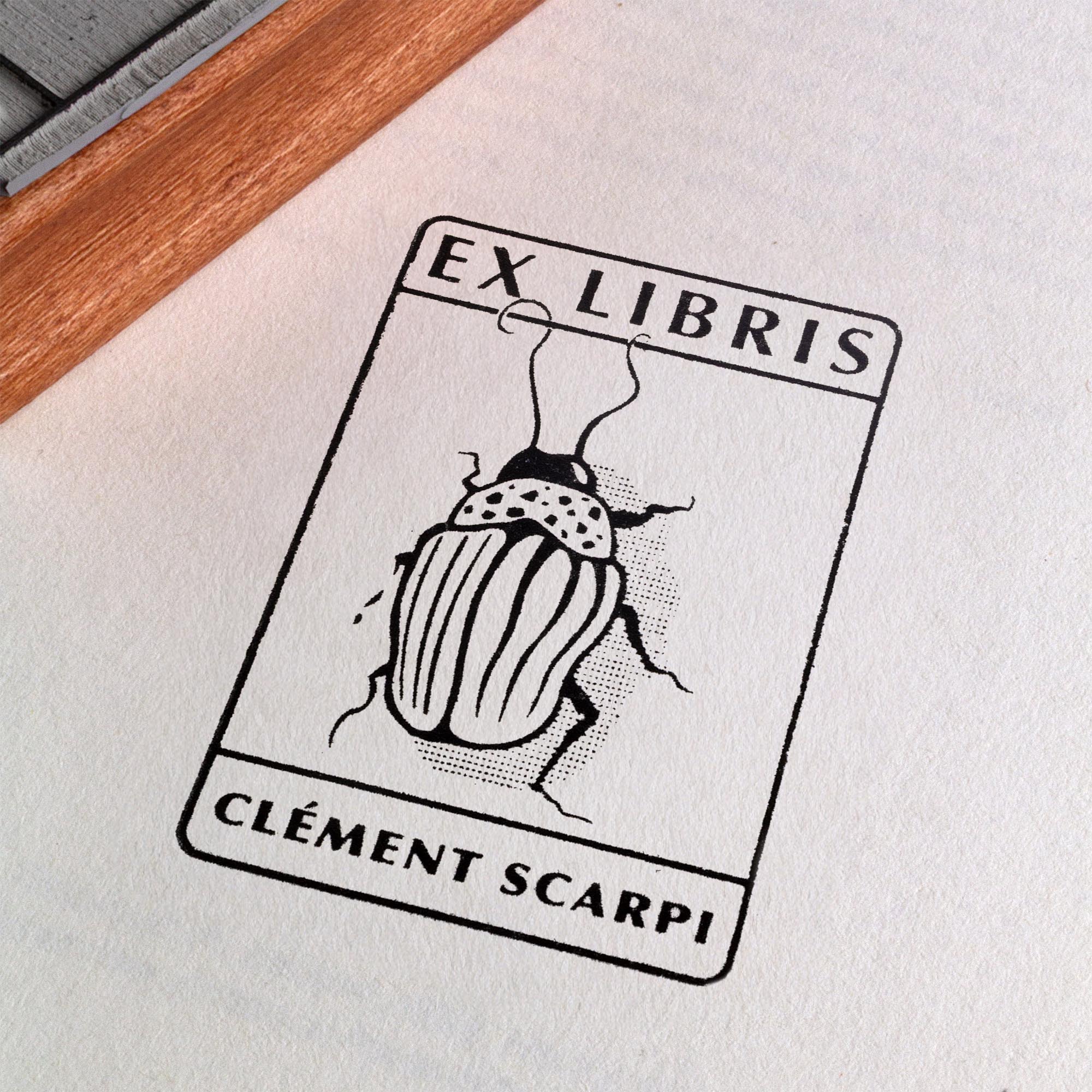 Tampon ex-libris personnalisé en bois et caoutchouc Insect I de Clément Scarpi. Accessoire de papeterie et de bibliothèque pour amoureux des livres représentant l'empreinte gravée d'un scarabée (insecte) sous l'inscription Ex Libris, posé à côté d'un encreur noir sur les pages ouvertes d'un livre.