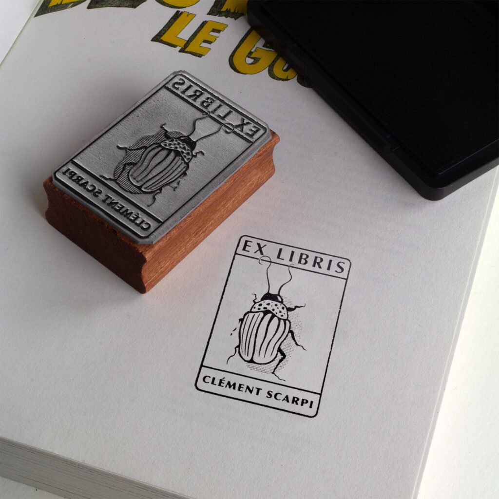 Tampon ex-libris personnalisé en bois et caoutchouc Insect I de Clément Scarpi. Accessoire de papeterie et de bibliothèque pour amoureux des livres représentant l'empreinte gravée d'un scarabée (insecte) sous l'inscription Ex Libris, posé à côté d'un encreur noir sur les pages ouvertes d'un livre.