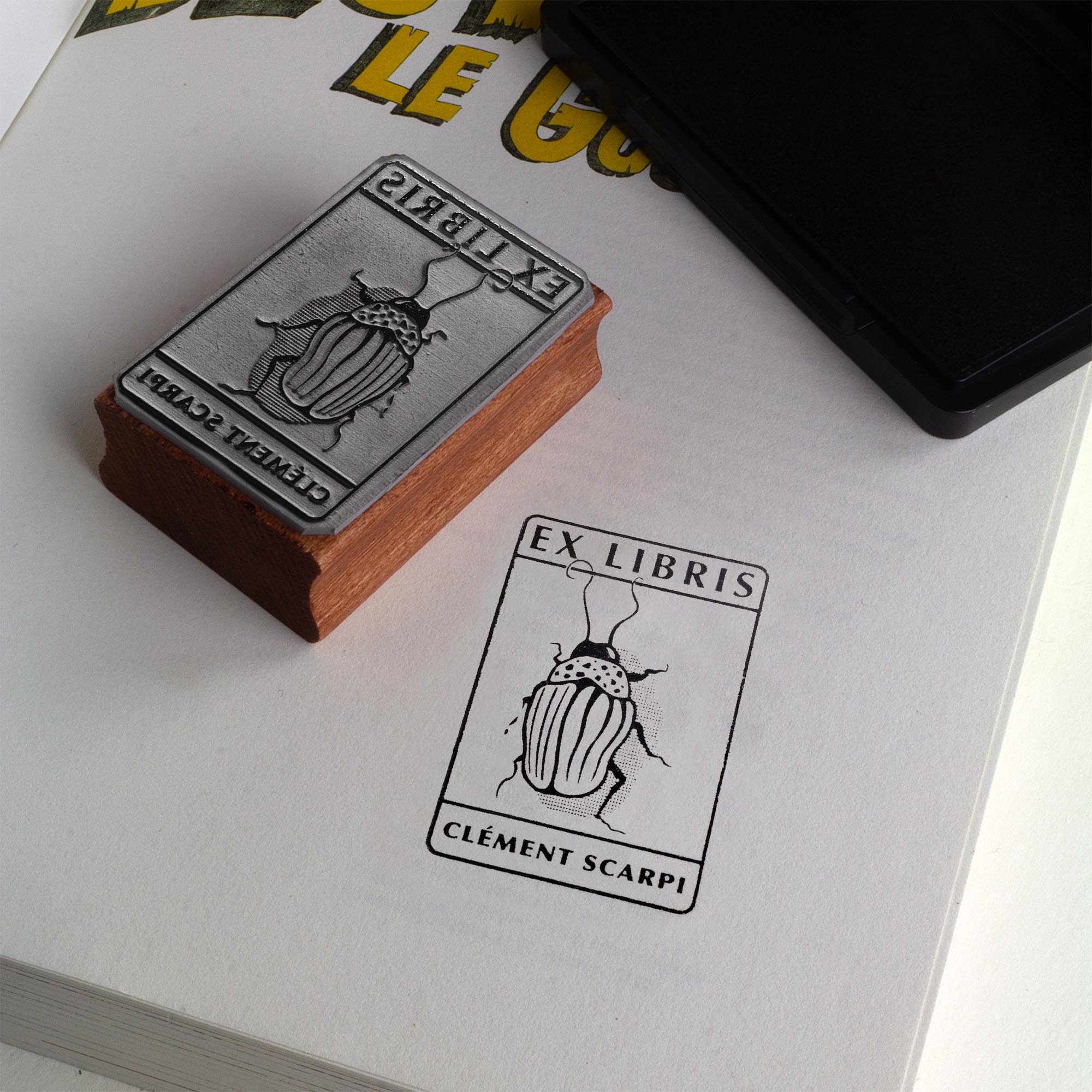 Tampon ex-libris personnalisé en bois et caoutchouc Insect I de Clément Scarpi. Accessoire de papeterie et de bibliothèque pour amoureux des livres représentant l'empreinte gravée d'un scarabée (insecte) sous l'inscription Ex Libris, posé à côté d'un encreur noir sur les pages ouvertes d'un livre.