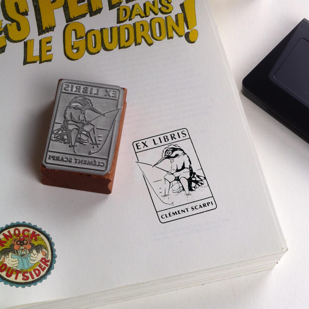 Tampon encreur Ex Libris Kingfisher de l'artiste Clément Scarpi, montrant le bloc en caoutchouc gravé à côté de son empreinte noire sur une page de livre, illustrant un martin-pêcheur humanisé qui pêche à la ligne