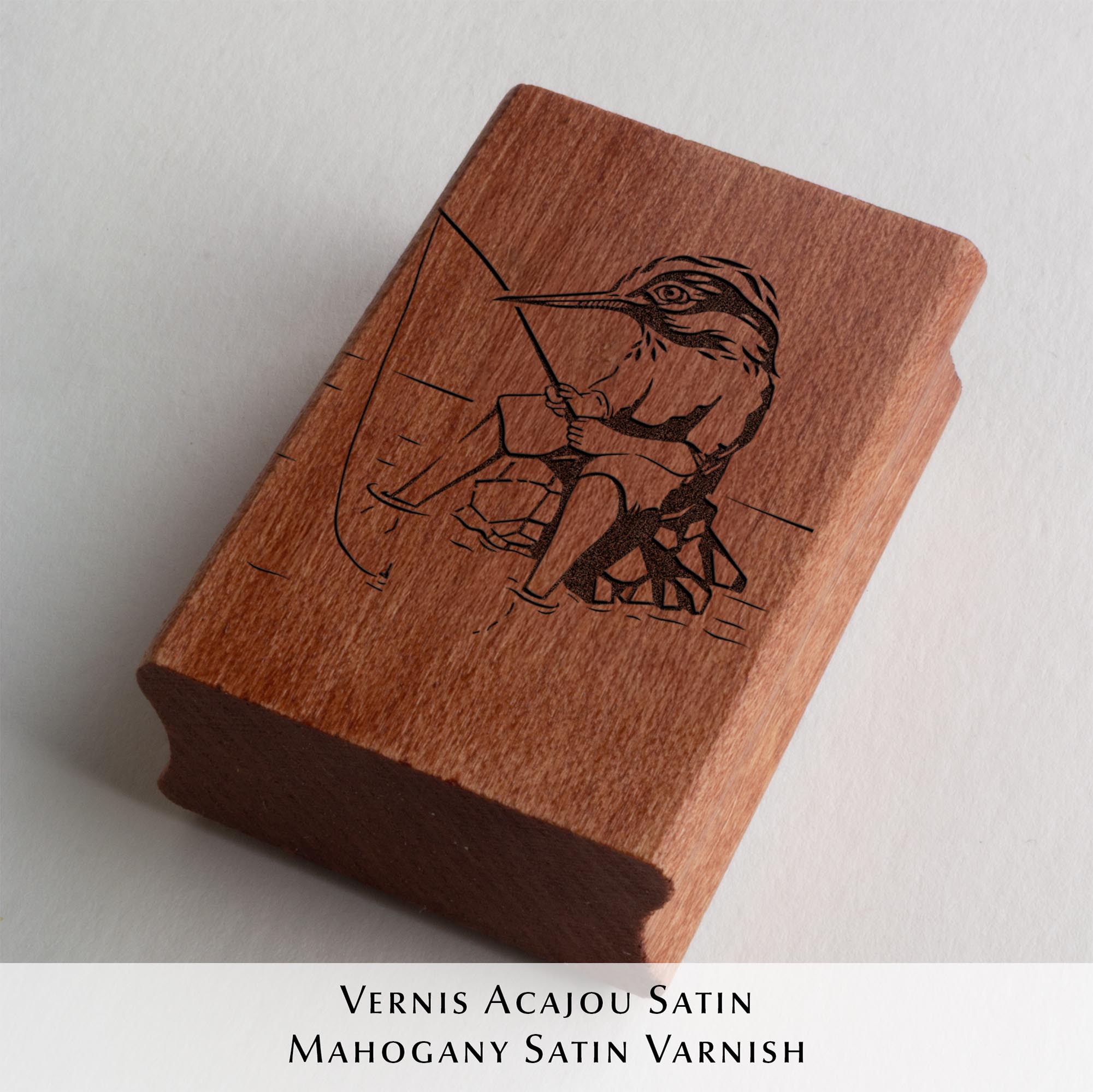Tampon encreur Ex Libris Kingfisher de l'artiste Clément Scarpi, montrant le bloc en caoutchouc gravé à côté de son empreinte noire sur une page de livre, illustrant un martin-pêcheur humanisé qui pêche à la ligne