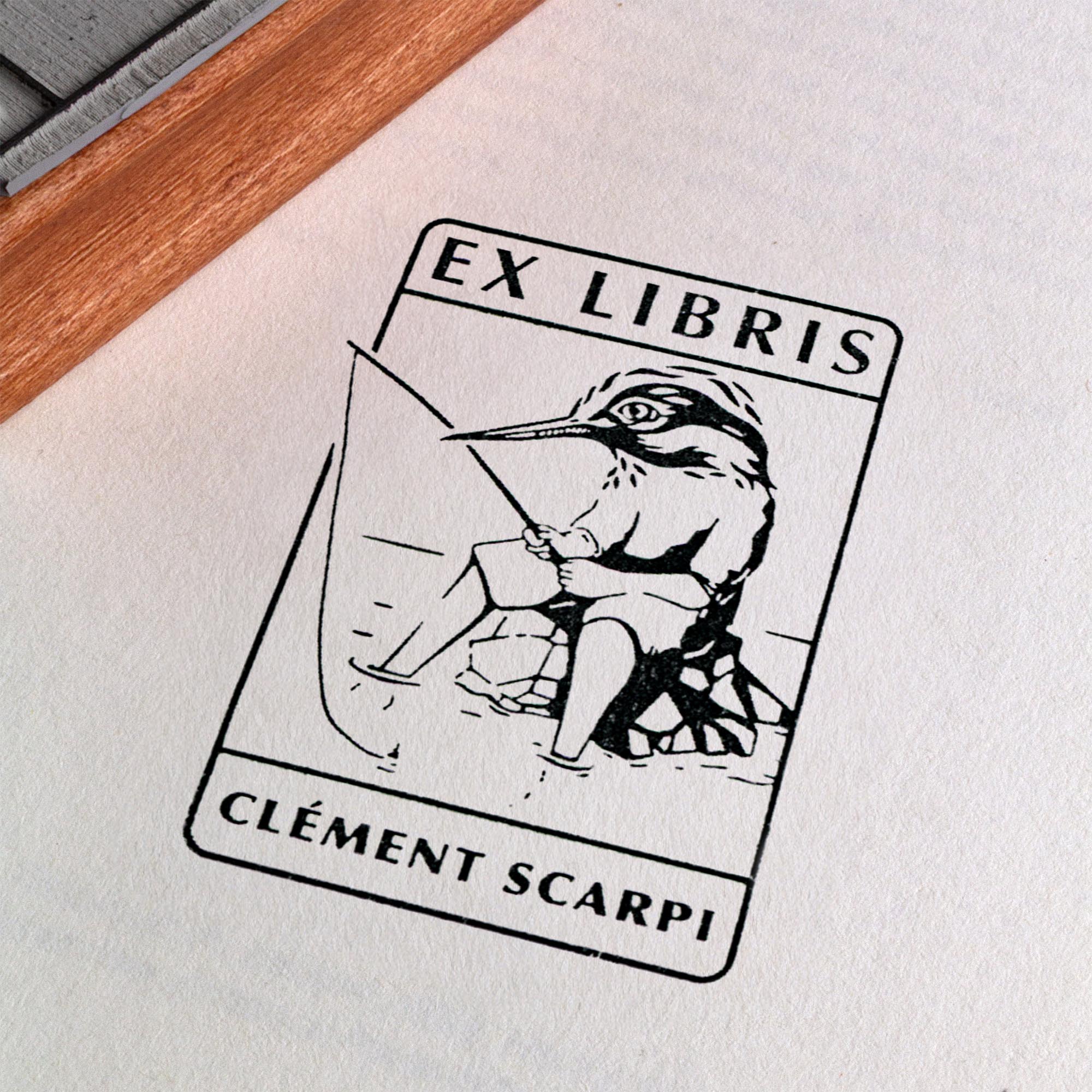 Tampon encreur Ex Libris Kingfisher de l'artiste Clément Scarpi, montrant le bloc en caoutchouc gravé à côté de son empreinte noire sur une page de livre, illustrant un martin-pêcheur humanisé qui pêche à la ligne