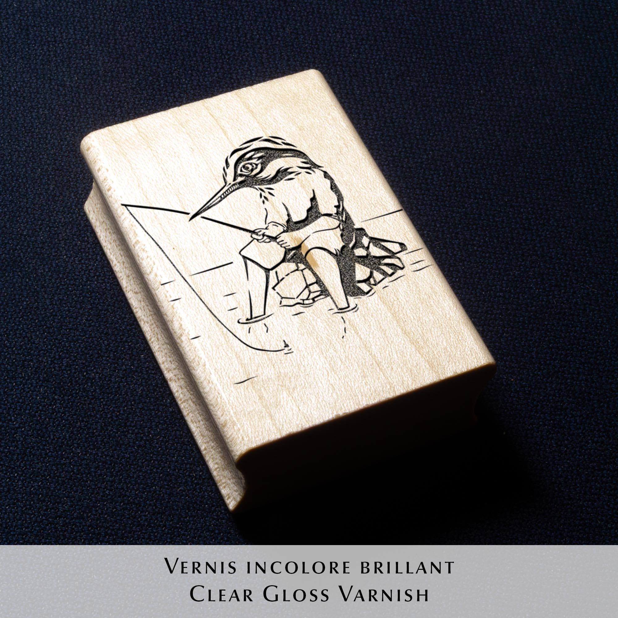 Tampon encreur Ex Libris Kingfisher de l'artiste Clément Scarpi, montrant le bloc en caoutchouc gravé à côté de son empreinte noire sur une page de livre, illustrant un martin-pêcheur humanisé qui pêche à la ligne