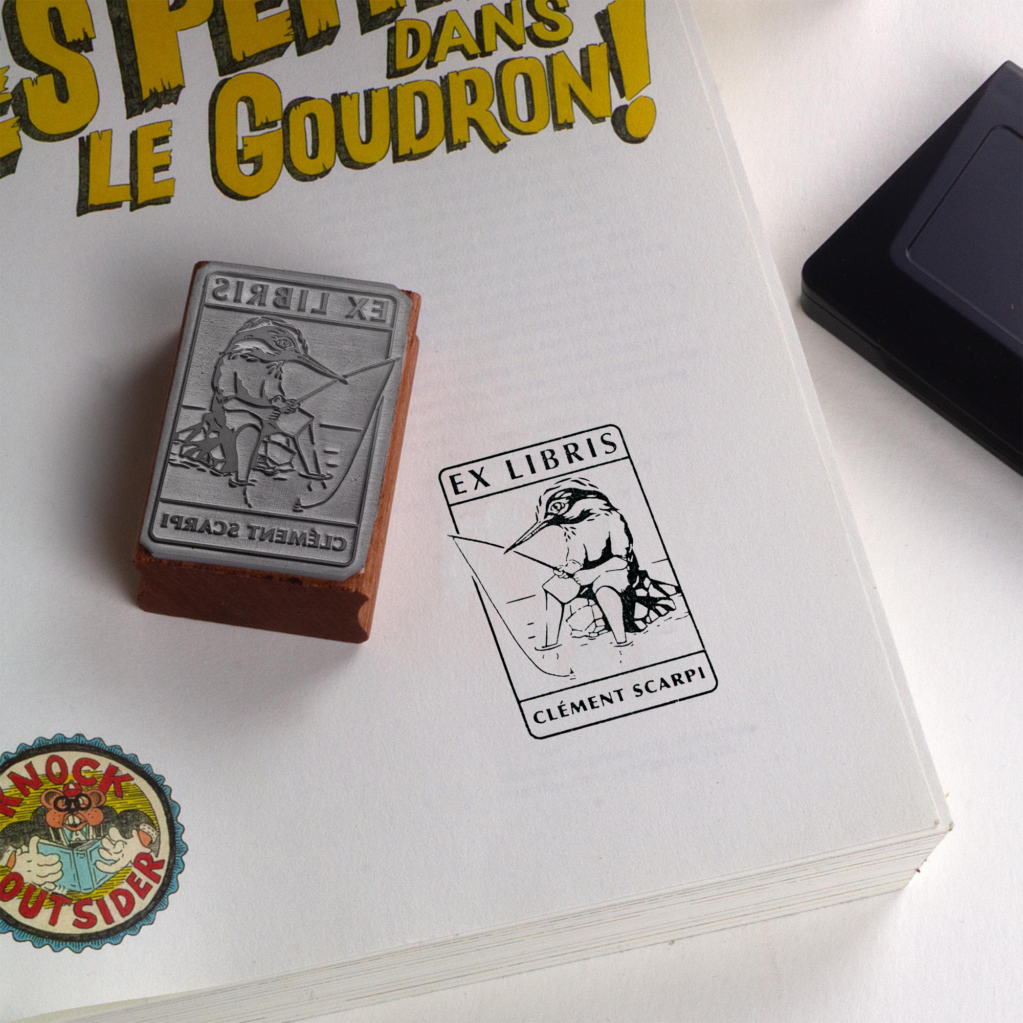 Tampon encreur Ex Libris Kingfisher de l'artiste Clément Scarpi, montrant le bloc en caoutchouc gravé à côté de son empreinte noire sur une page de livre, illustrant un martin-pêcheur humanisé qui pêche à la ligne