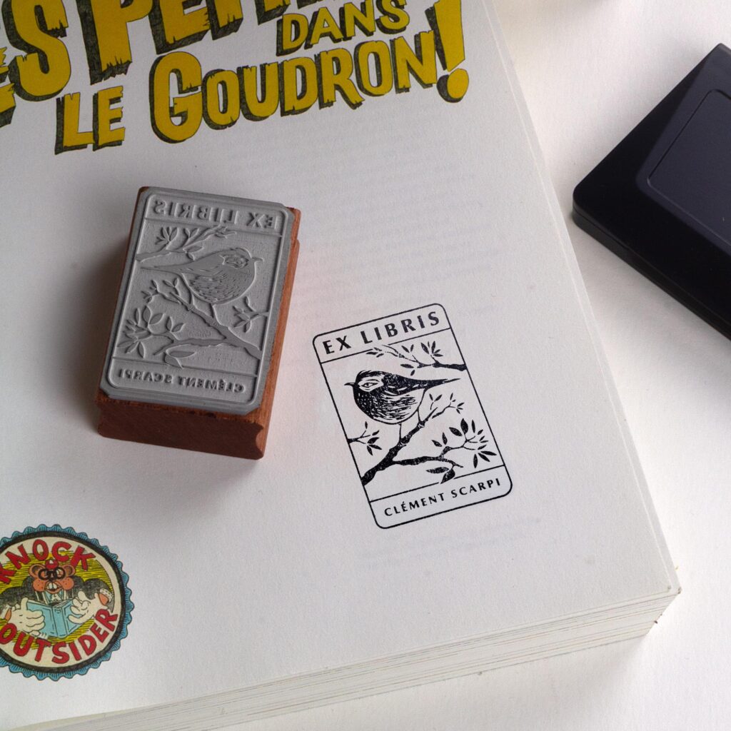 Tampon ex-libris en bois et caoutchouc The Kinglet de Clément Scarpi. Marque-page et accessoire de bibliothèque personnalisé représentant une empreinte de gravure de roitelet (oiseau) sur une branche avec l'inscription Ex Libris, posé sur les pages ouvertes d'un livre.