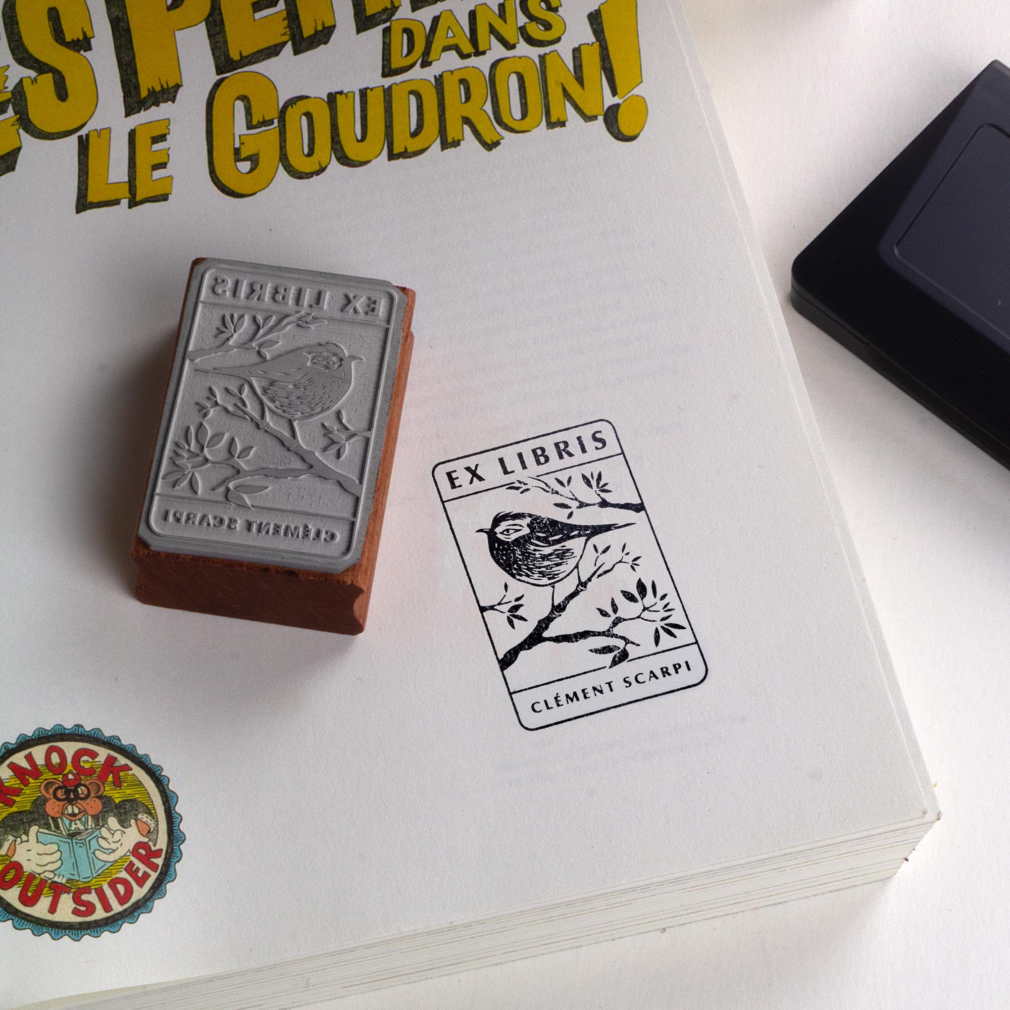Tampon ex-libris en bois et caoutchouc The Kinglet de Clément Scarpi. Marque-page et accessoire de bibliothèque personnalisé représentant une empreinte de gravure de roitelet (oiseau) sur une branche avec l'inscription Ex Libris, posé sur les pages ouvertes d'un livre.