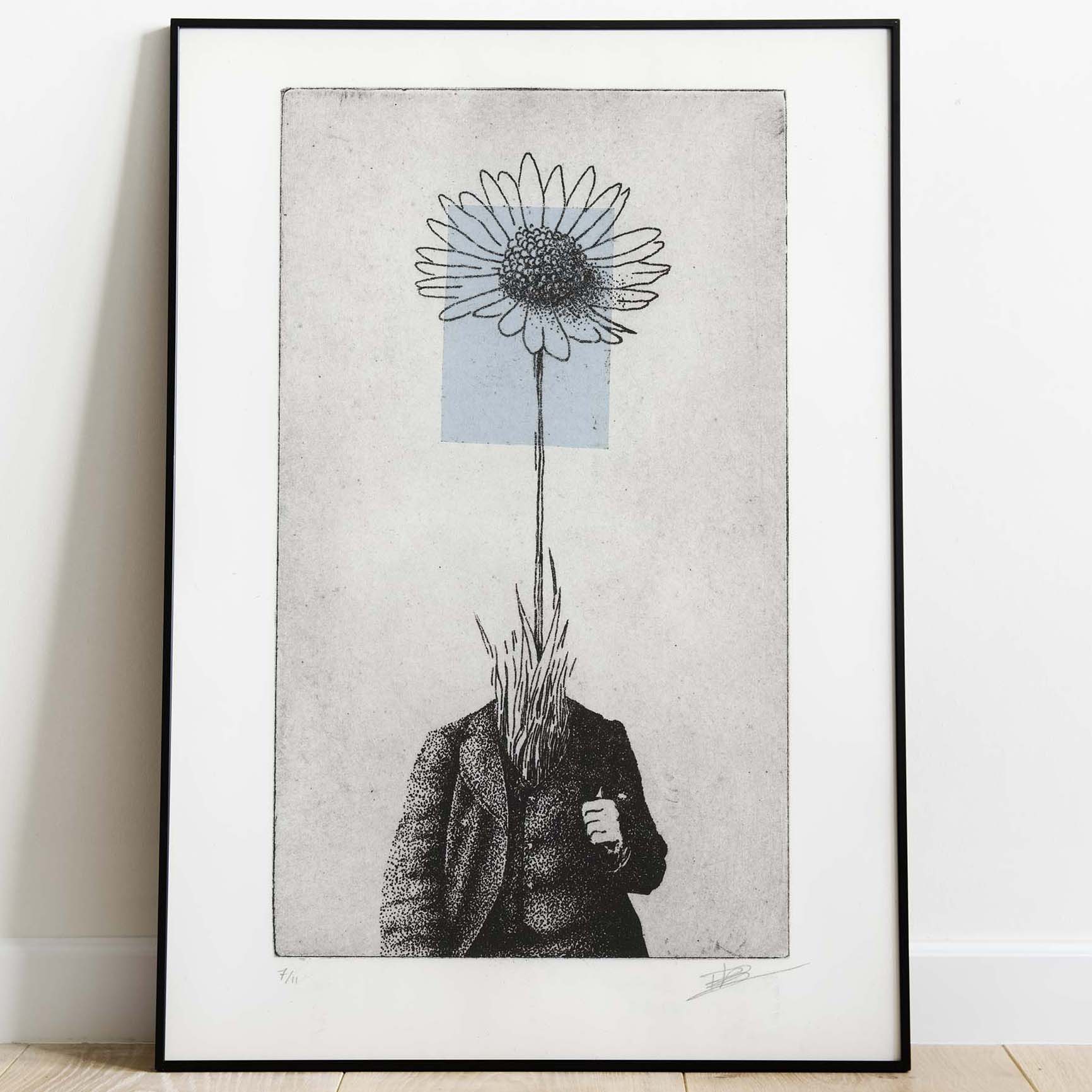 Gravure surréaliste "Bellis perennis" dans un grand cadre noir fin, posée au sol sur un parquet en bois clair contre un mur blanc. L'œuvre verticale montre un personnage en costume dont la tête est remplacée par une grande marguerite sur fond bleu.