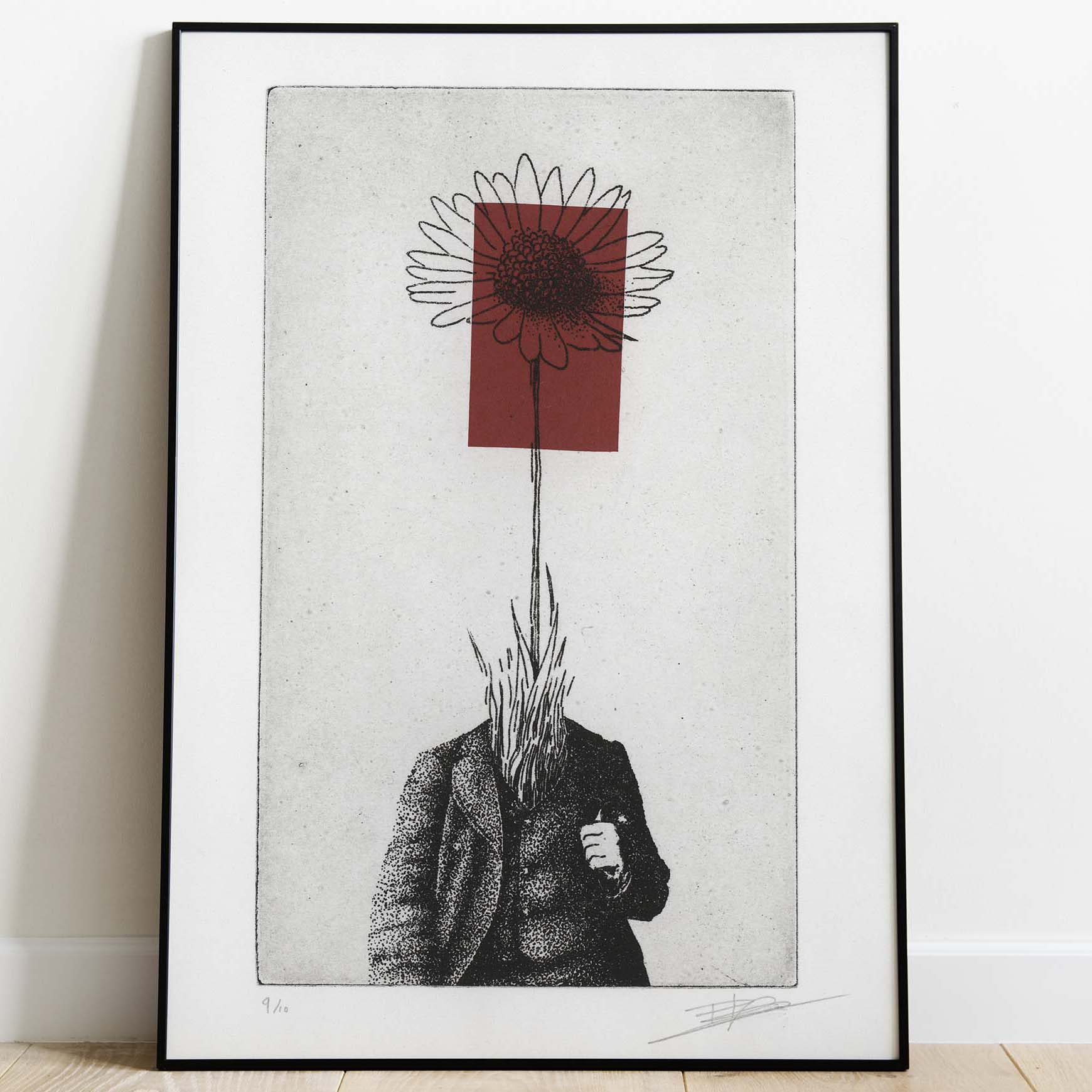 Gravure surréaliste "Bellis perennis" dans un grand cadre noir fin, posée au sol sur un parquet en bois clair contre un mur blanc. L'œuvre verticale montre un personnage en costume dont la tête est remplacée par une grande marguerite sur fond rouge.