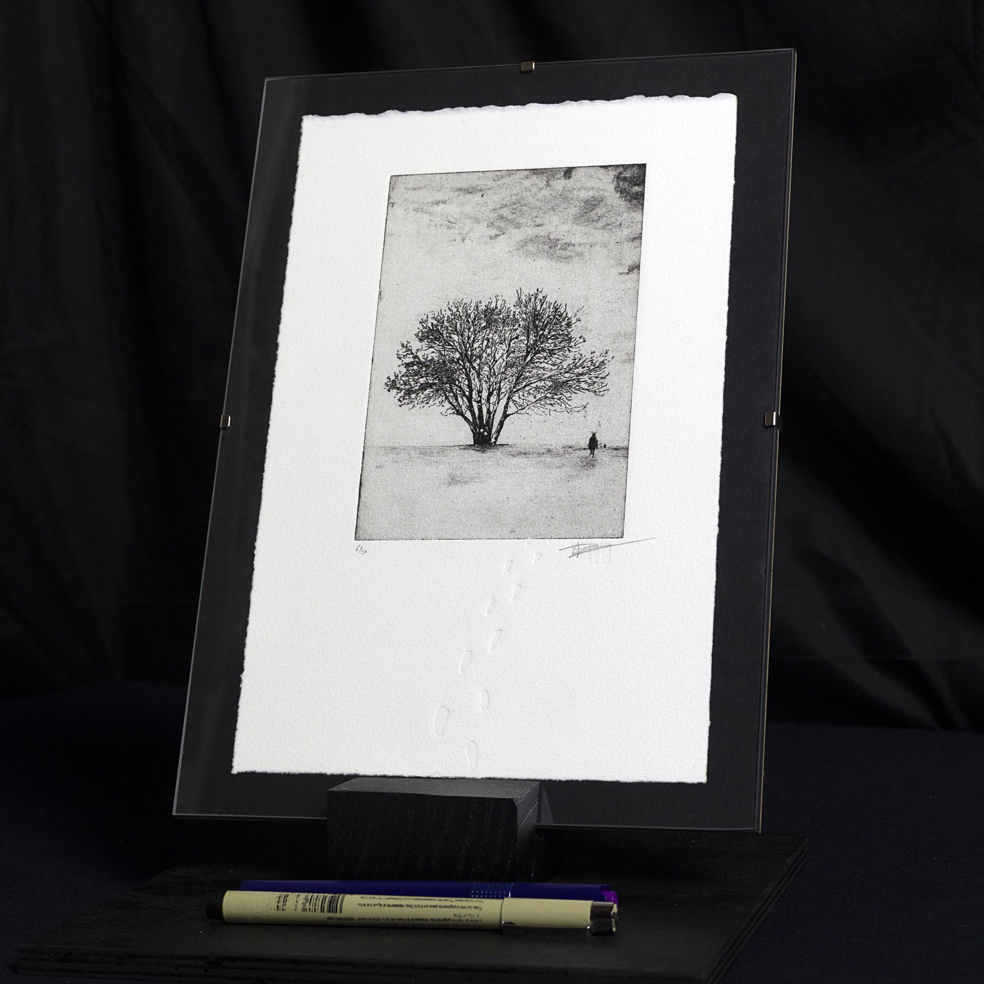 Gravure originale Footprints in the snow de Clément Scarpi. Estampe d'art en noir et blanc représentant un paysage d'hiver enneigé avec un cycliste au loin et des traces de pas en relief (gaufrage) sur papier d'art Velin Arches BFK Rives.
