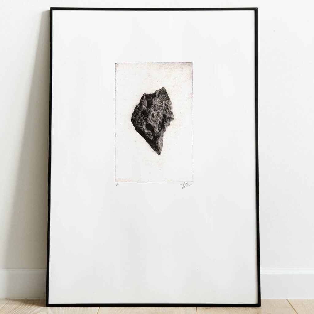 Estampe originale texturée intitulée "Caillou I". L'illustration à l'encre noire affiche un personnage de profil, dont le buste et l'épaule sont suggérés par des hachures et des aplats sombres. Sa tête et son cou se métamorphosent en un bloc de pierre ou un gros caillou à la texture minérale fortement détaillée, créant une silhouette poétique et mystérieuse. L'ensemble est imprimé sur une feuille de papier d'art blanc épais aux contours irréguliers faits main. En bas de la feuille, sous la cuve d'impression, apparaissent la numérotation du tirage limité à gauche et la signature manuscrite de l'artiste à droite. Le papier est photographié à plat sur un fond noir contrasté.