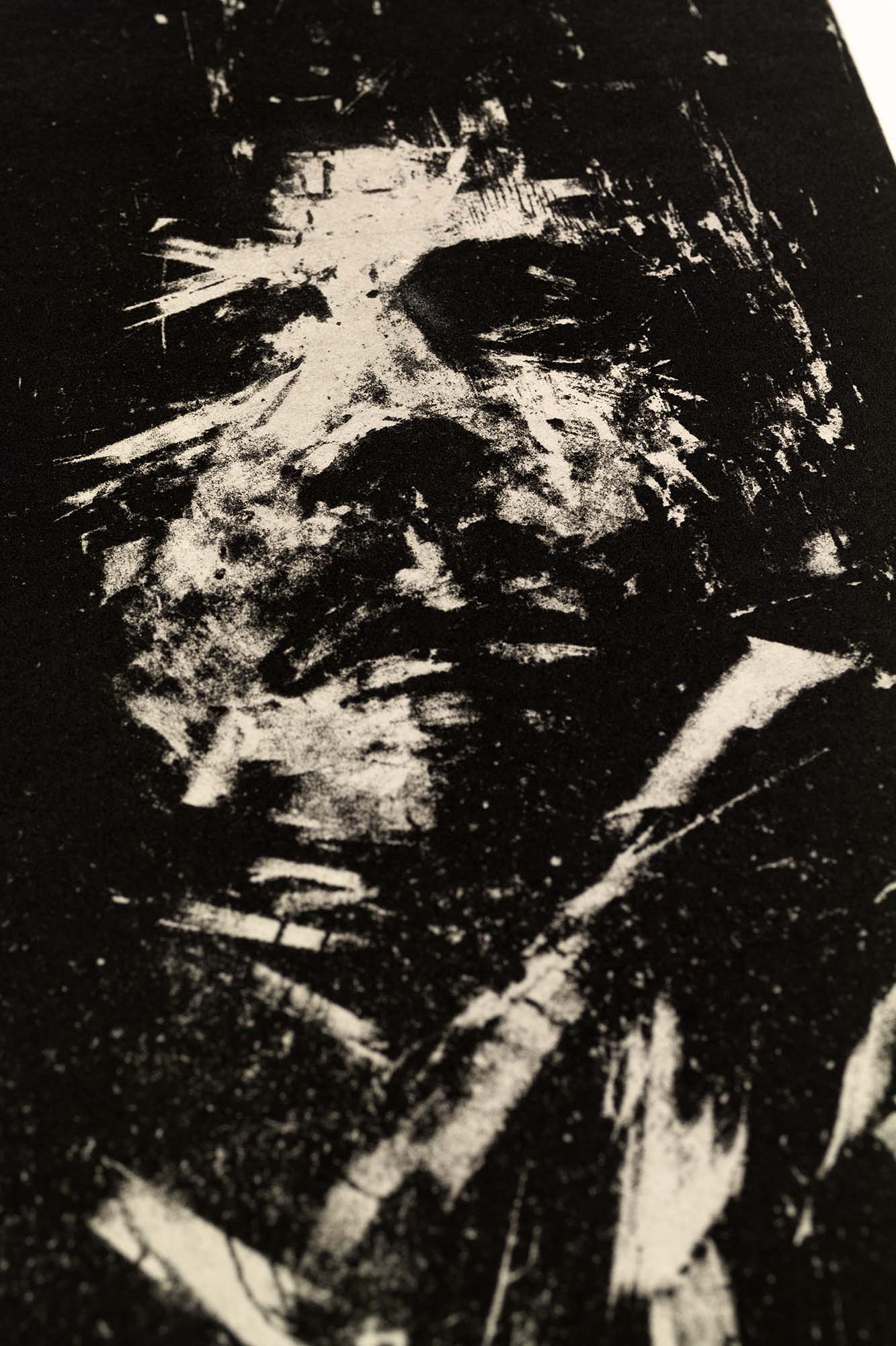 Gravure expressionniste en noir et blanc "Portrait" dans un grand cadre noir fin avec un passe-partout blanc, posée au sol sur un parquet clair contre un mur blanc.