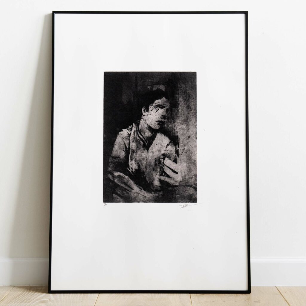 Mise en situation d'un grand cadre noir rectangulaire posé sur un sol en parquet de bois clair contre un mur blanc. À l'intérieur, un large passe-partout blanc met en valeur une estampe verticale à l'encre noire. L'illustration montre un visage masculin fragmenté et ténébreux, émergeant de touches d'encre texturées et de lignes gravées à l'esthétique brute et mélancolique. Sous la gravure, on distingue les inscriptions au crayon « EA » à gauche et la signature de l'artiste à droite.