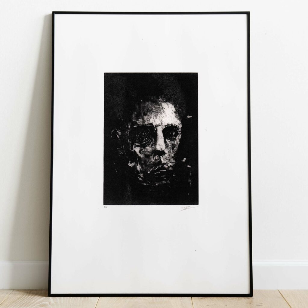 Mise en situation d'un grand cadre noir rectangulaire posé sur un sol en parquet de bois clair contre un mur blanc. À l'intérieur, un large passe-partout blanc met en valeur une estampe verticale à l'encre noire. L'illustration montre un visage masculin fragmenté et ténébreux, émergeant de touches d'encre texturées et de lignes gravées à l'esthétique brute et mélancolique. Sous la gravure, on distingue les inscriptions au crayon « EA » à gauche et la signature de l'artiste à droite.