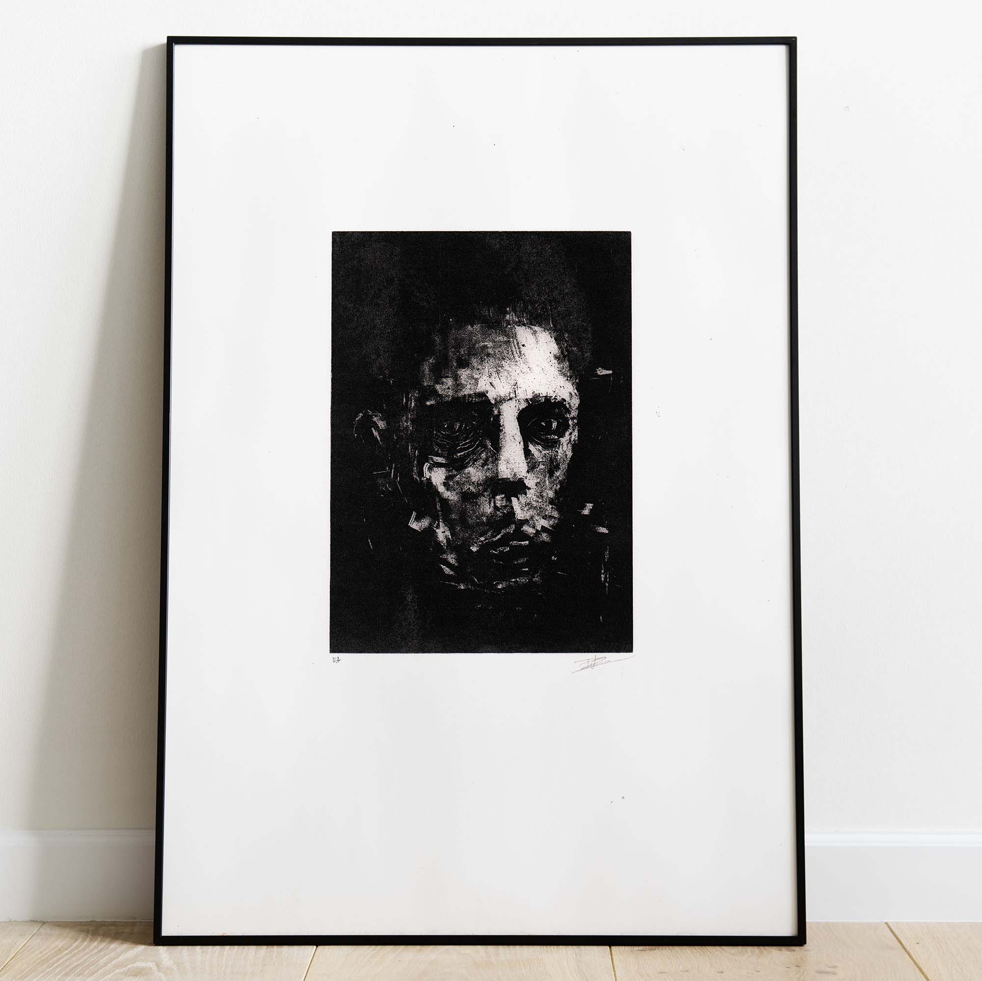 Mise en situation d'un grand cadre noir rectangulaire posé sur un sol en parquet de bois clair contre un mur blanc. À l'intérieur, un large passe-partout blanc met en valeur une estampe verticale à l'encre noire. L'illustration montre un visage masculin fragmenté et ténébreux, émergeant de touches d'encre texturées et de lignes gravées à l'esthétique brute et mélancolique. Sous la gravure, on distingue les inscriptions au crayon « EA » à gauche et la signature de l'artiste à droite.