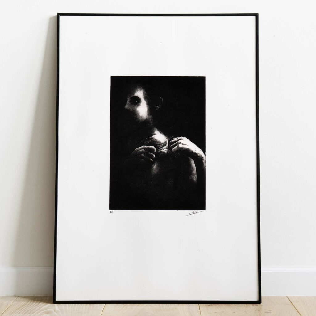 Mise en situation d'un grand cadre noir rectangulaire posé sur un sol en parquet de bois clair contre un mur blanc. À l'intérieur, un large passe-partout blanc met en valeur une estampe verticale à l'encre noire. L'illustration montre un visage masculin fragmenté et ténébreux, émergeant de touches d'encre texturées et de lignes gravées à l'esthétique brute et mélancolique. Sous la gravure, on distingue les inscriptions au crayon « EA » à gauche et la signature de l'artiste à droite.