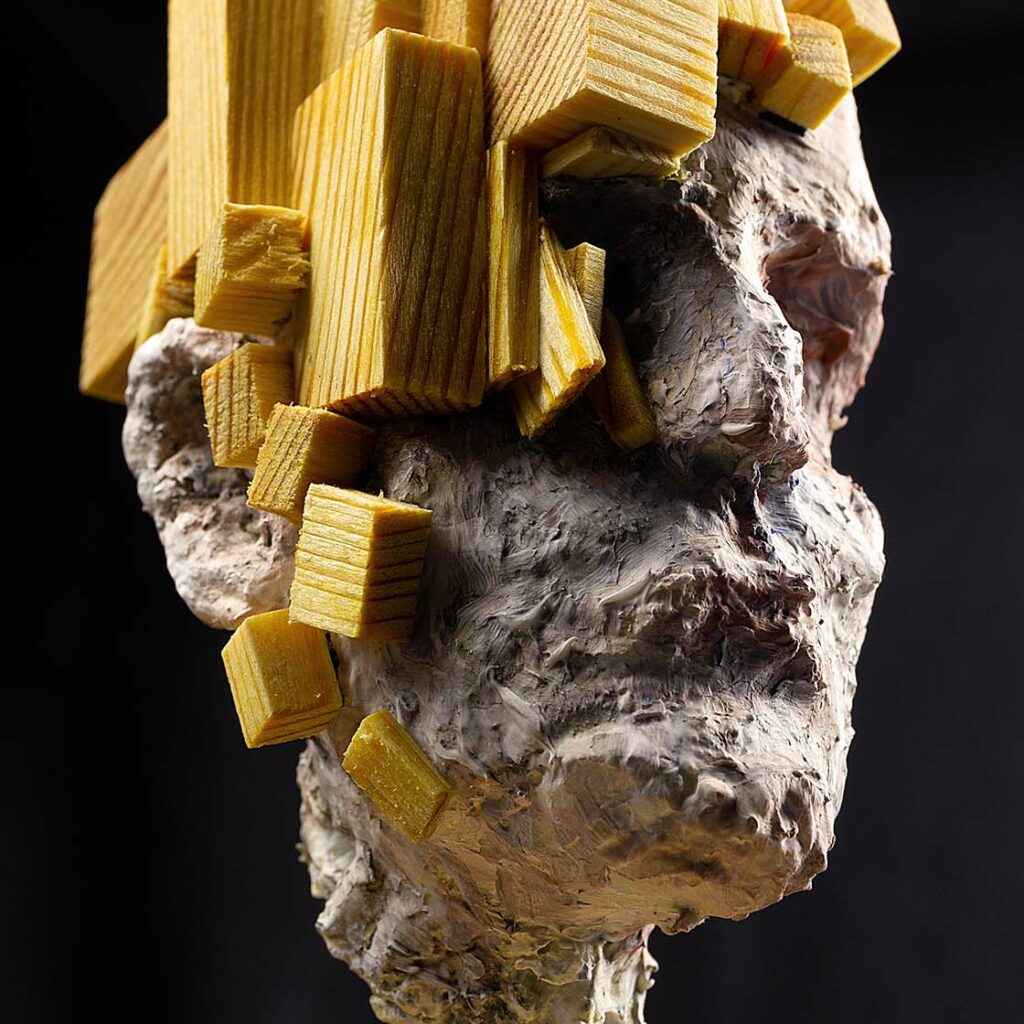 Sculpture contemporaine "Head I" représentant un visage en papier mâché texturé blanc, surmonté et partiellement recouvert de blocs de bois géométriques jaunes, fixée sur une tige métallique verticale reliée à un socle circulaire en bois clair.